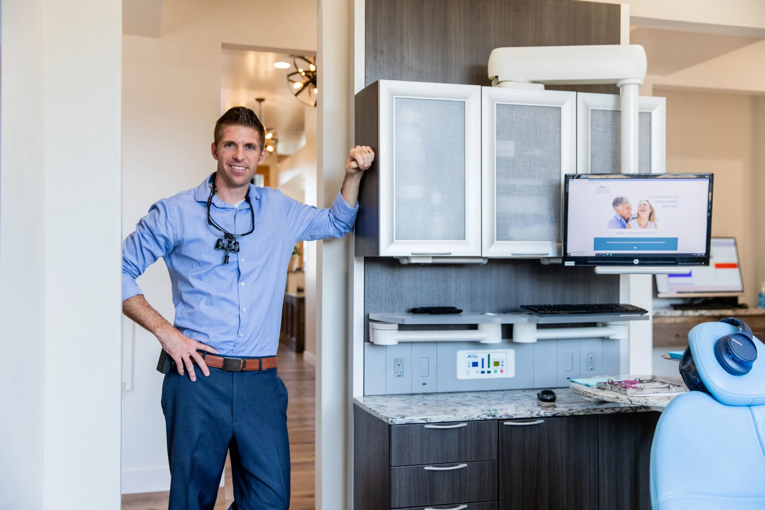 A smiling dentist in Twin Falls, dressed in business attire, stands in a modern dental office next to cabinets, with dental tools and a blue chair visible. A computer screen displays a website featuring a happy couple.