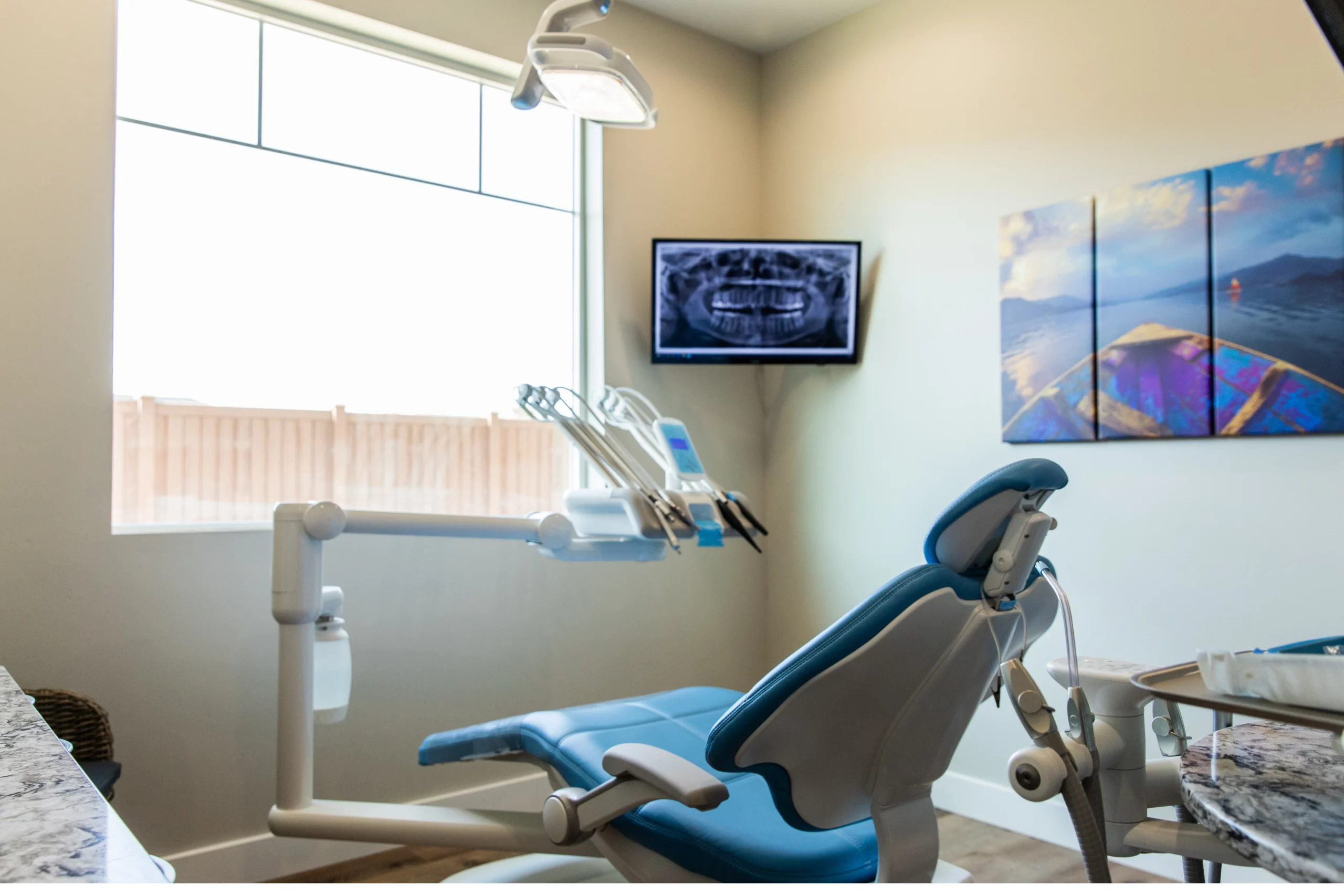 A modern dental exam room with a blue dental chair, overhead light, dental instruments, and a wall-mounted screen displaying a dental X-ray. Large window and wall art create a bright, welcoming atmosphere for patients visiting the dentist in Twin Falls.