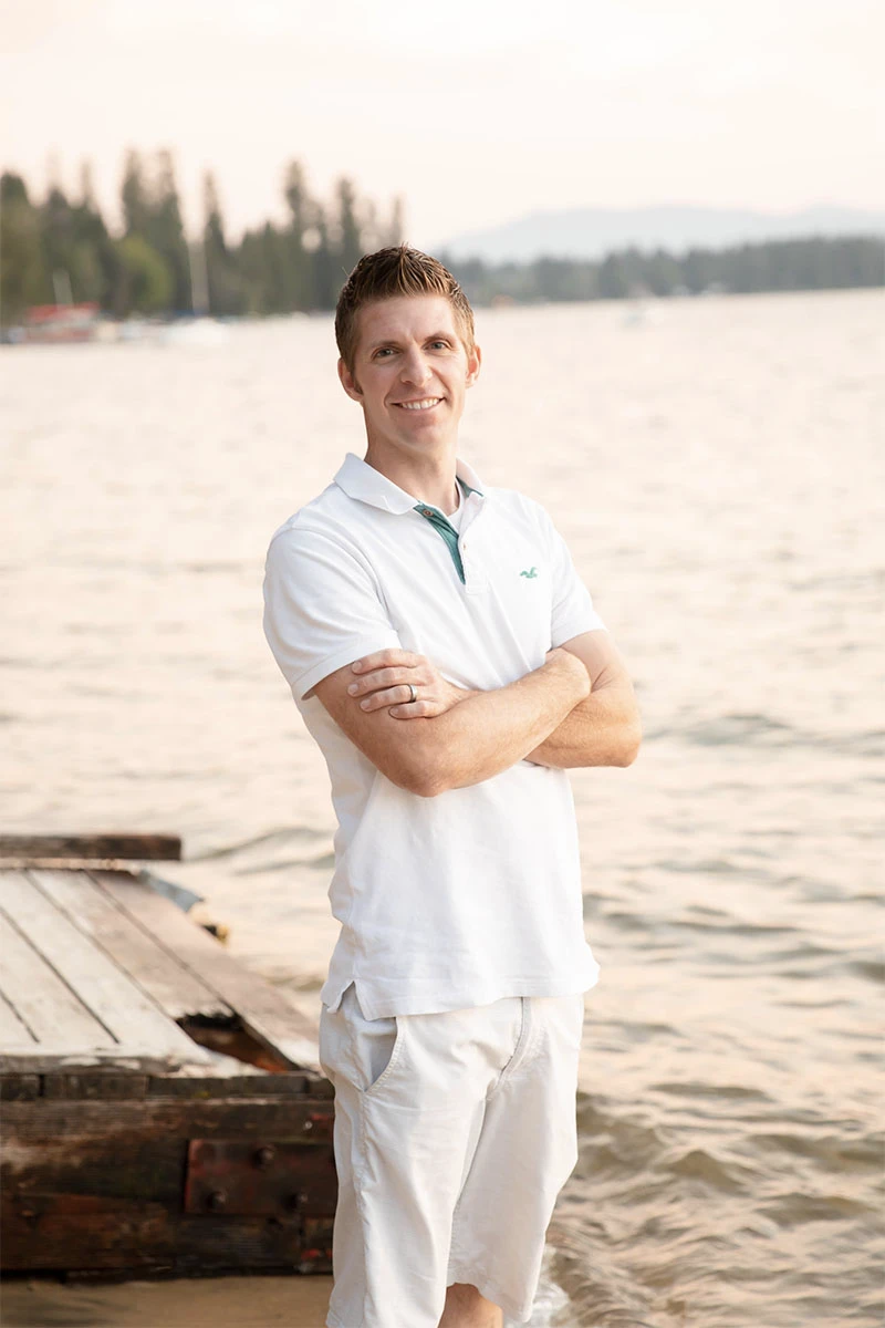 A dentist in Twin Falls, wearing a white polo shirt and light shorts, stands with arms crossed on a wooden dock by a calm lake, with trees and mountains in the background on a sunny day.